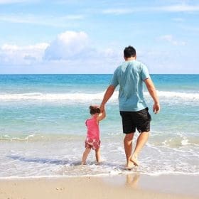 Pai segurando a mão de sua filha na praia, caminhando juntos , simbolizando a conexão e o amor no Dia dos Pais